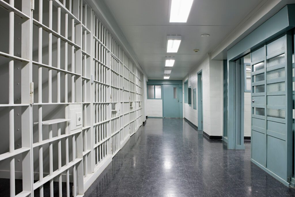 Empty correctional facility hallway with barred jail cells, secured doors, and overhead fluorescent lighting.
